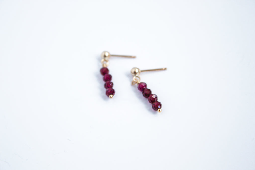 Moment and Milestone stud earrings in 14k gold filled and dangling garnet gemstones, symbolizing life’s meaningful moments, displayed on a white background.