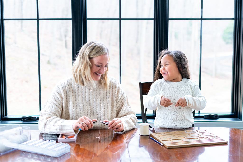 Olivia and Becky, the owner, making a stretch bracelet at a kitchen table.