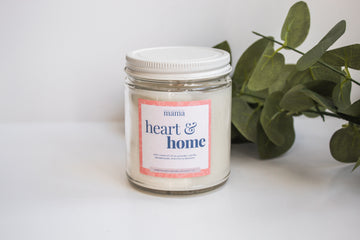 Heart and home branded candle with soft lighting and greenery background.